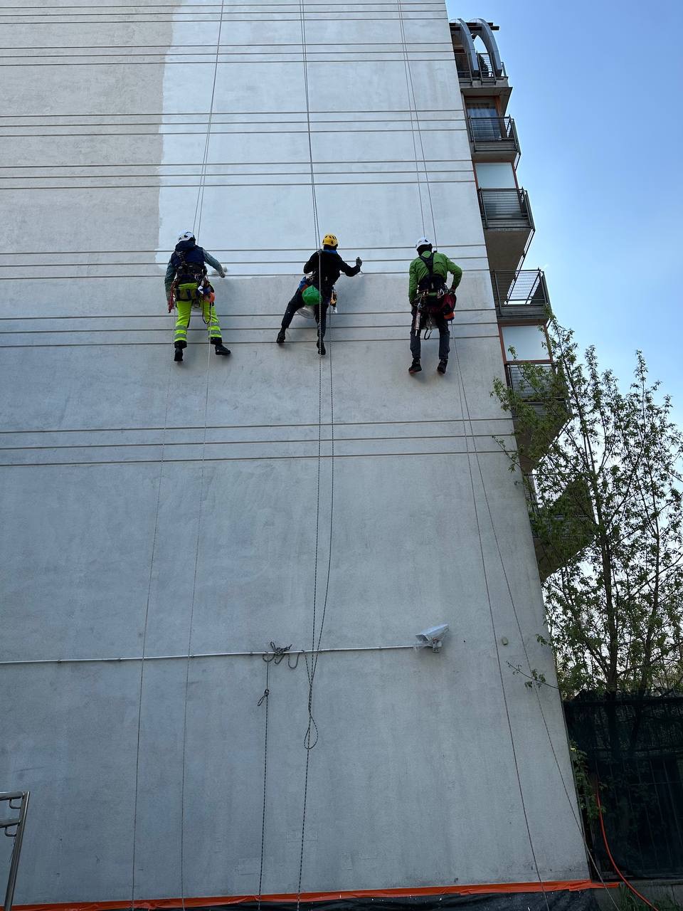 Trzech alpinistów przemysłowych maluje elewację wysokiego budynku. Widoczny fragment pomalowanej ściany, liny, zabezpieczenia i sprzęt alpinistyczny.