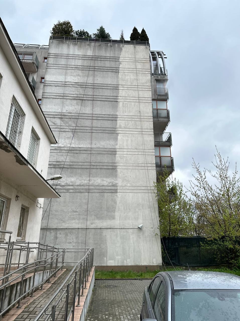 Częściowo umyta, szara elewacja wysokiego budynku z balkonami, widoczna z dołu, z fragmentem drugiego budynku po lewej i samochodem w prawym dolnym rogu.