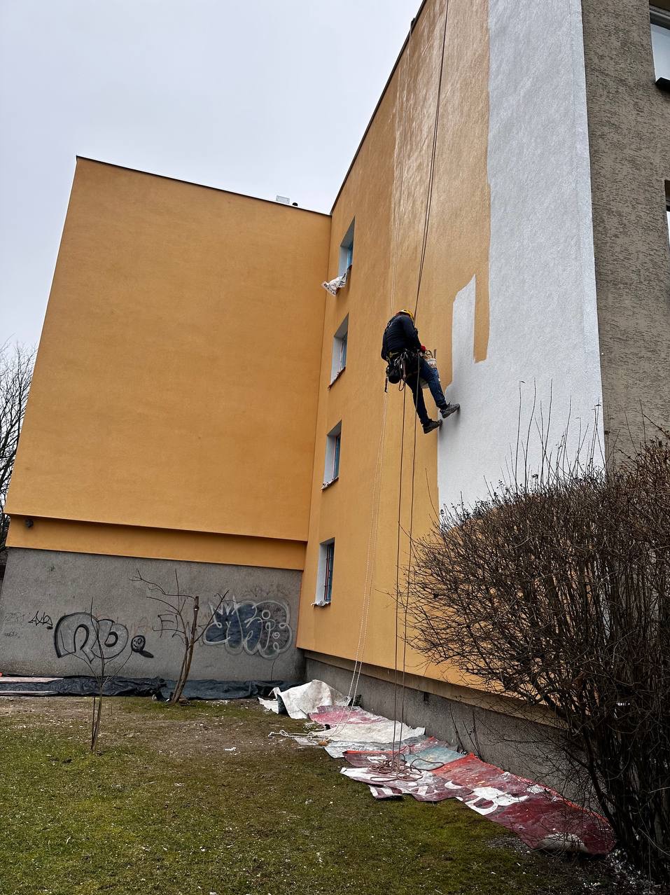 Pracownik na linach maluje fragment elewacji budynku na biało. Część budynku w kolorze żółtym, widoczne graffiti na cokole. Rozłożone zabezpieczenia na trawie.