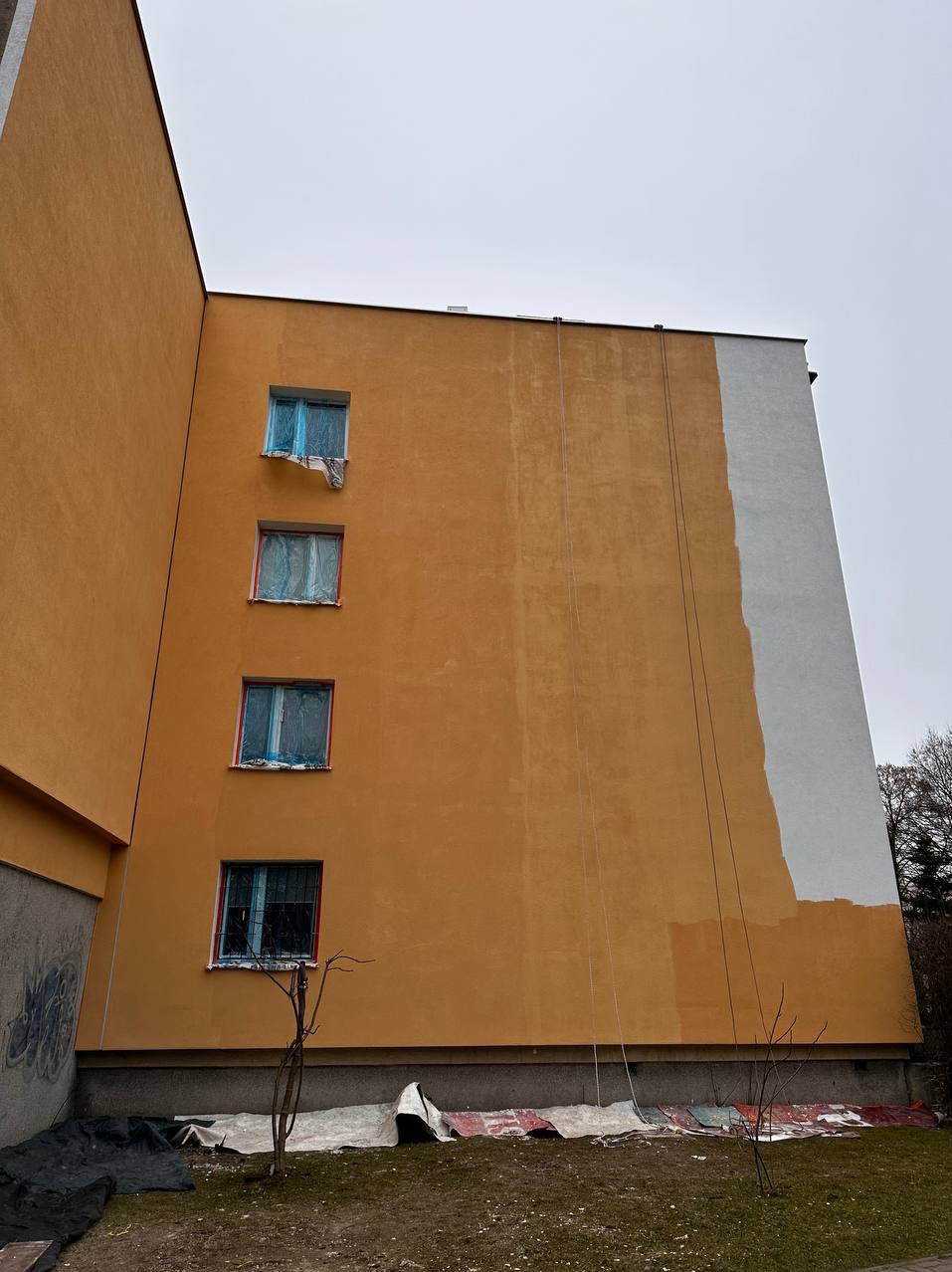 Żółta elewacja bloku z trzema oknami zabezpieczonymi folią, fragment pomalowany na biało. Widoczne liny i zabezpieczenia na ziemi. Ujęcie z dołu, pochmurne niebo.