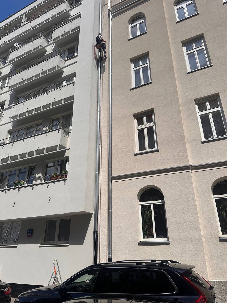 Pracownik na linie, w trakcie prac na elewacji budynku z jasną fasadą. Widoczne okna i elementy architektoniczne. U dołu zaparkowany samochód.