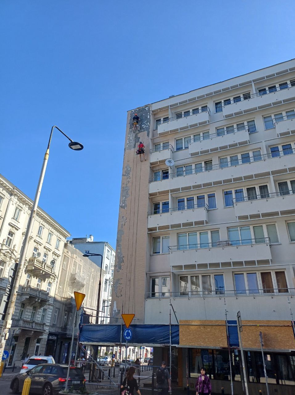 Dwóch alpinistów na linach odnawia elewację wysokiego budynku w mieście. Widoczne rusztowania i fragmenty odnowionej ściany. Błękitne niebo w tle.