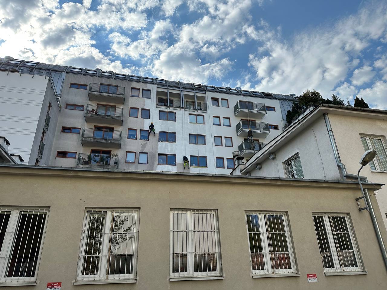 Dwóch pracowników na linach zabezpieczających czyści elewację wysokiego budynku. Część ściany jest już umyta, ukazując różnicę w kolorze. Błękitne niebo z chmurami w tle.