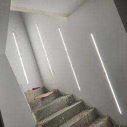 Instalacje elektryczne Piotr Pater - Schody w trakcie remontu z zamontowanym oświetleniem liniowym w ścianie. Widoczne narzędzia i przewody. Surowe wykończenie betonu kontrastuje z nowoczesnym światłem.