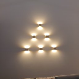 Instalacje elektryczne Piotr Pater - Symetryczny układ siedmiu kinkietów na ścianie w Krakowie, tworzących trójkątny wzór. Ciepłe światło oświetla gładką powierzchnię. Widoczne zabezpieczenia na dole ściany.