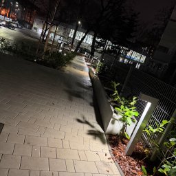 Instalacje elektryczne Piotr Pater - Nocne oświetlenie ścieżki z kostki brukowej, z lampami wzdłuż ogrodzenia i roślinności. Widok na budynek w tle, oświetlony ulicznymi latarniami.