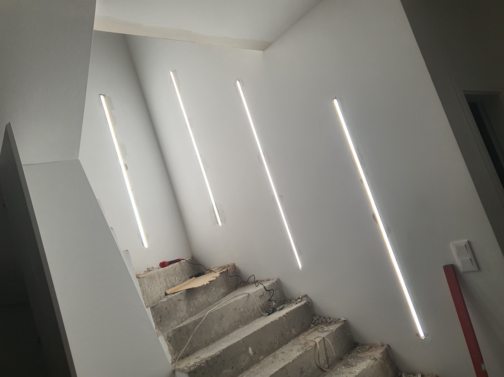 Schody w trakcie remontu z zamontowanym oświetleniem liniowym w ścianie. Widoczne narzędzia i przewody. Surowe wykończenie betonu kontrastuje z nowoczesnym światłem.