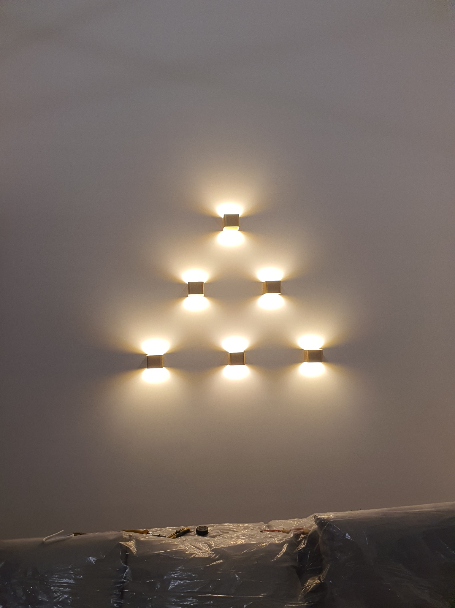 Symetryczny układ siedmiu kinkietów na ścianie w Krakowie, tworzących trójkątny wzór. Ciepłe światło oświetla gładką powierzchnię. Widoczne zabezpieczenia na dole ściany.