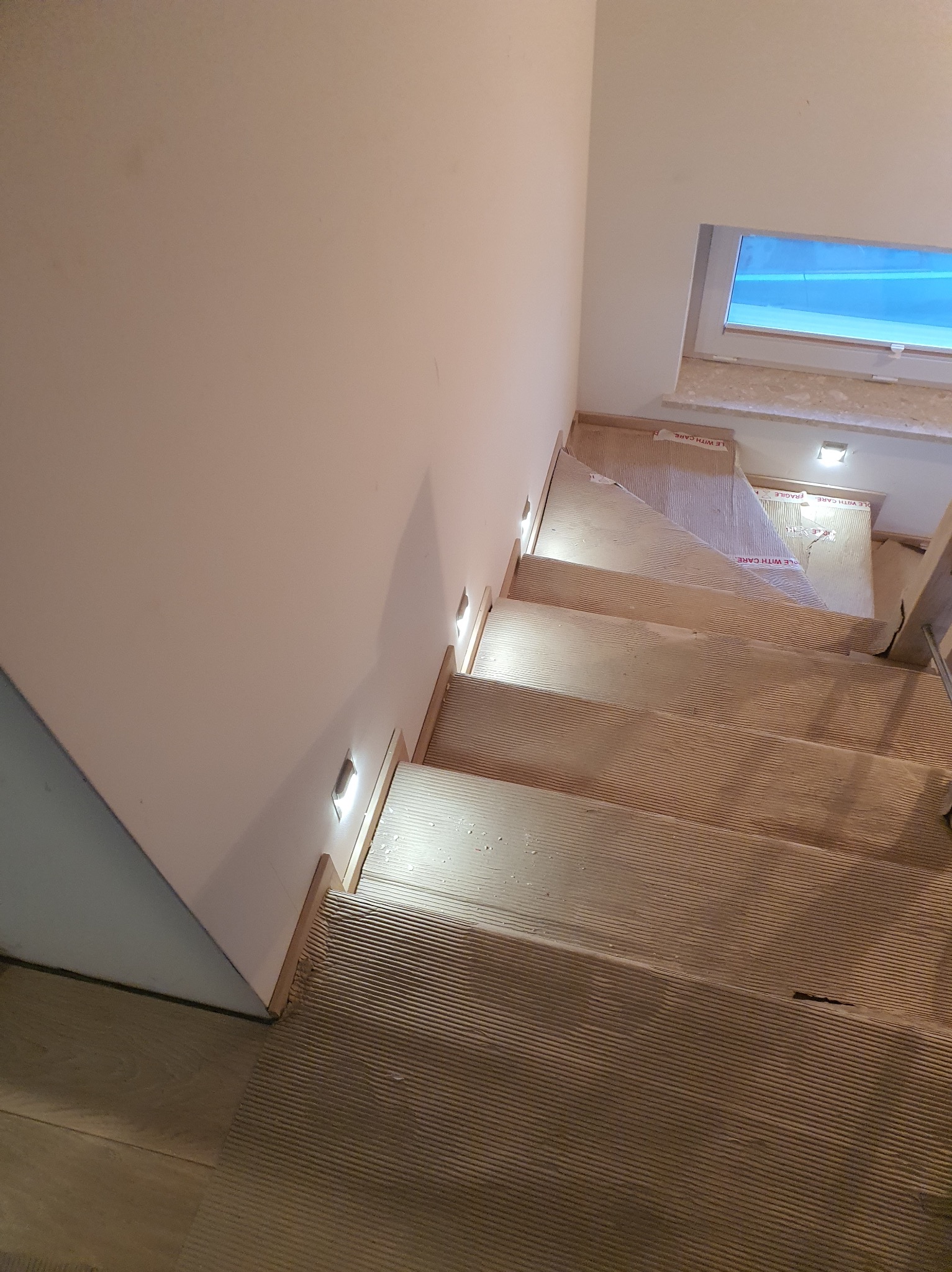 Schody z podświetleniem LED w ścianie, widok z góry. Stopnie zabezpieczone kartonem. Nowoczesne oświetlenie schodowe.