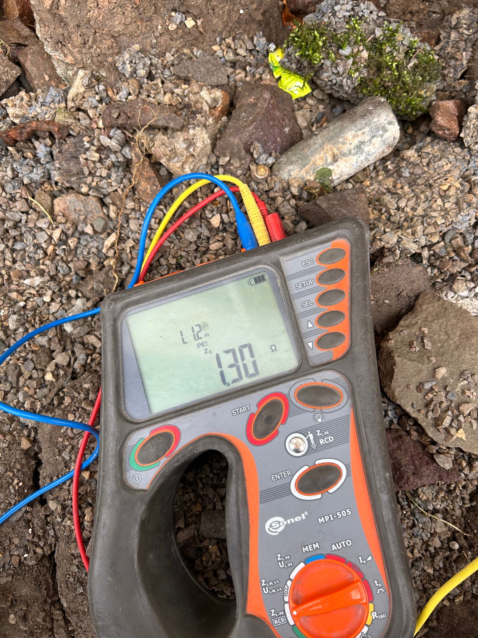 Miernik Sonel MPI-505 z podłączonymi przewodami pomiarowymi na tle ziemi i kamieni, wskazujący wartość 1.30 Ω, używany podczas pomiarów.