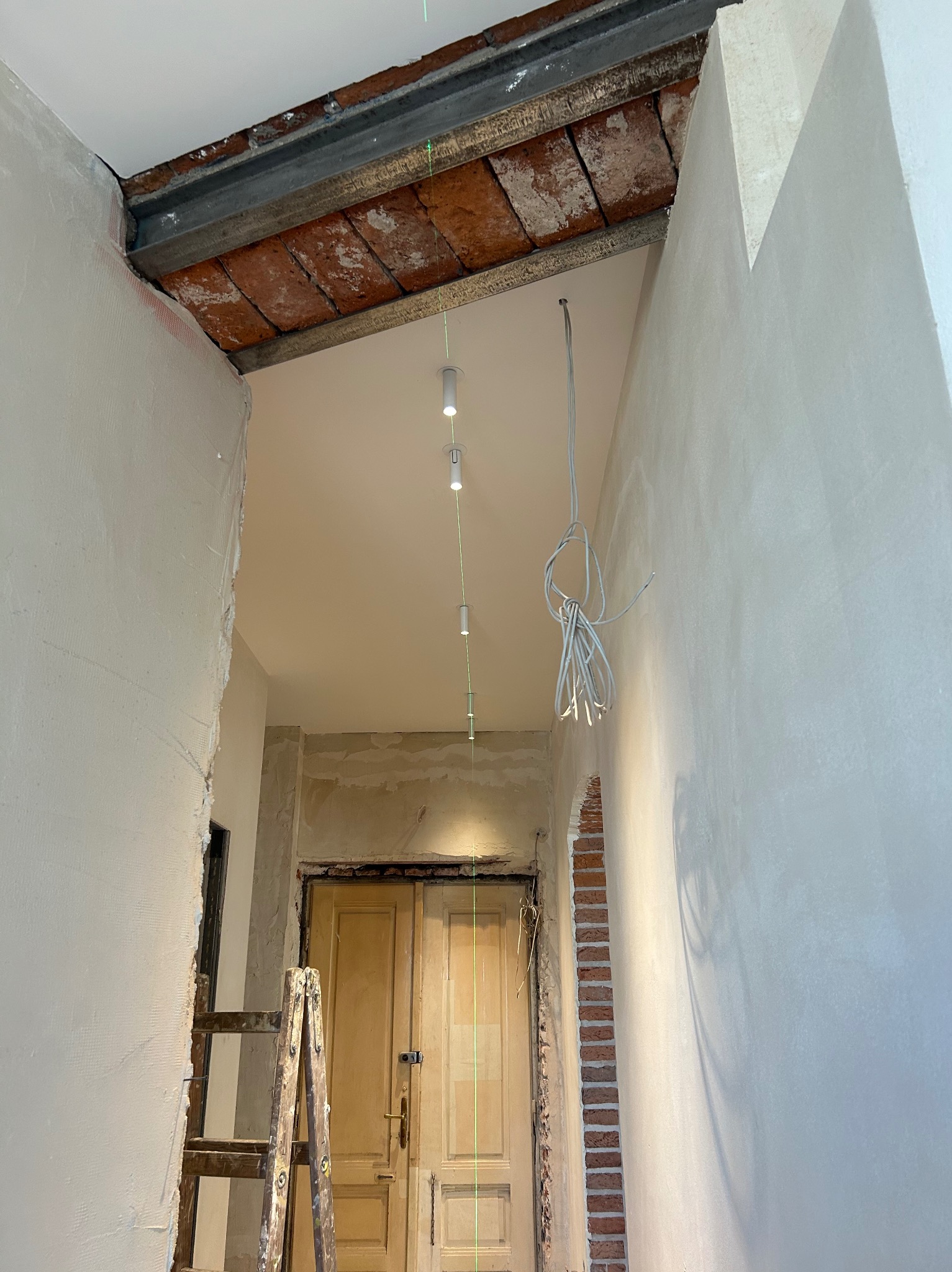 Wnętrze w trakcie remontu: widoczne przewody, zamontowane oświetlenie punktowe, surowa ściana z cegły, drabina i laserowy wskaźnik wyznaczający linię.
