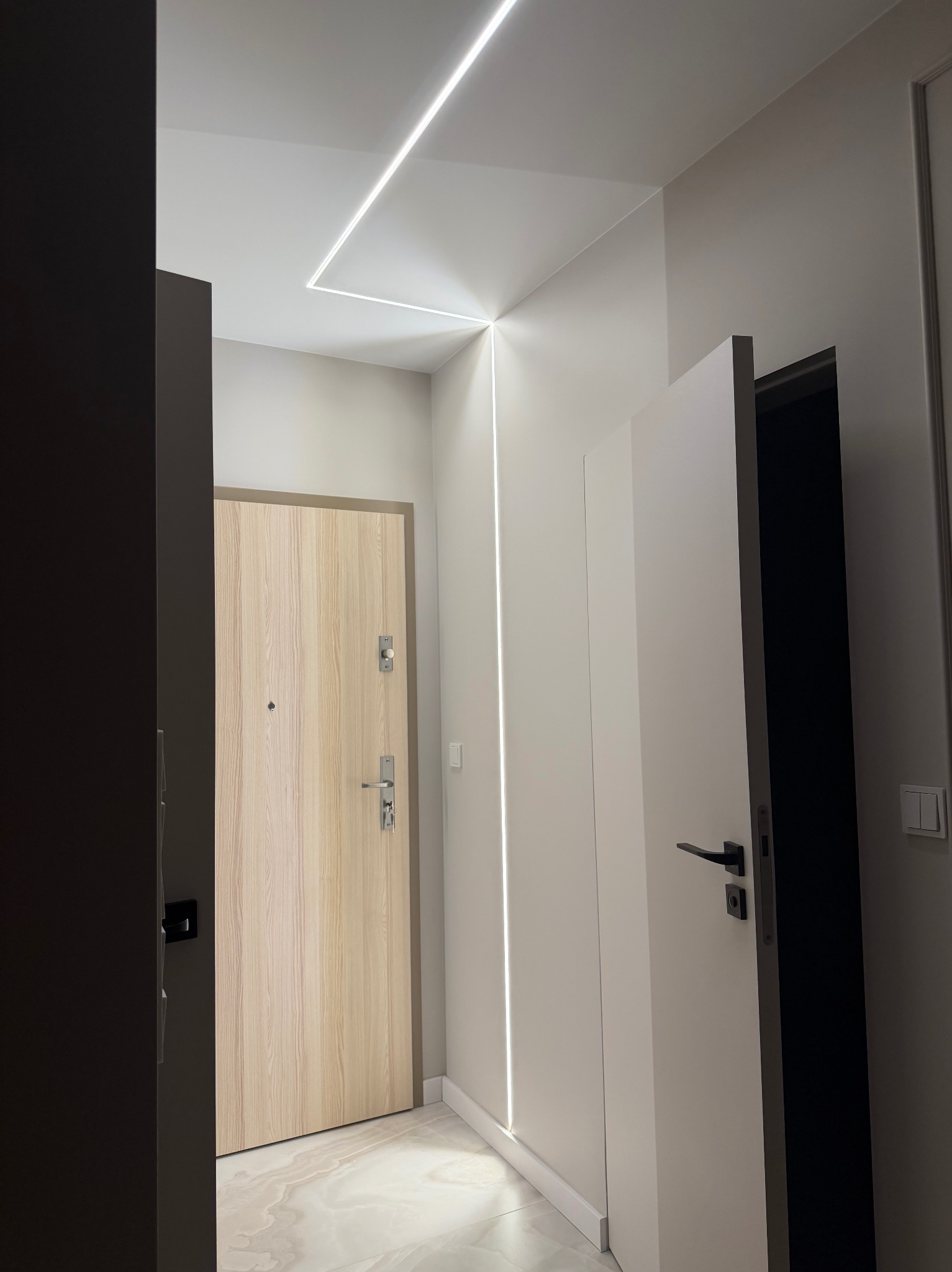 Nowoczesny przedpokój z oświetleniem LED w suficie i ścianie, tworzącym geometryczny wzór. Widoczne drzwi wejściowe i wewnętrzne, marmurowa podłoga.