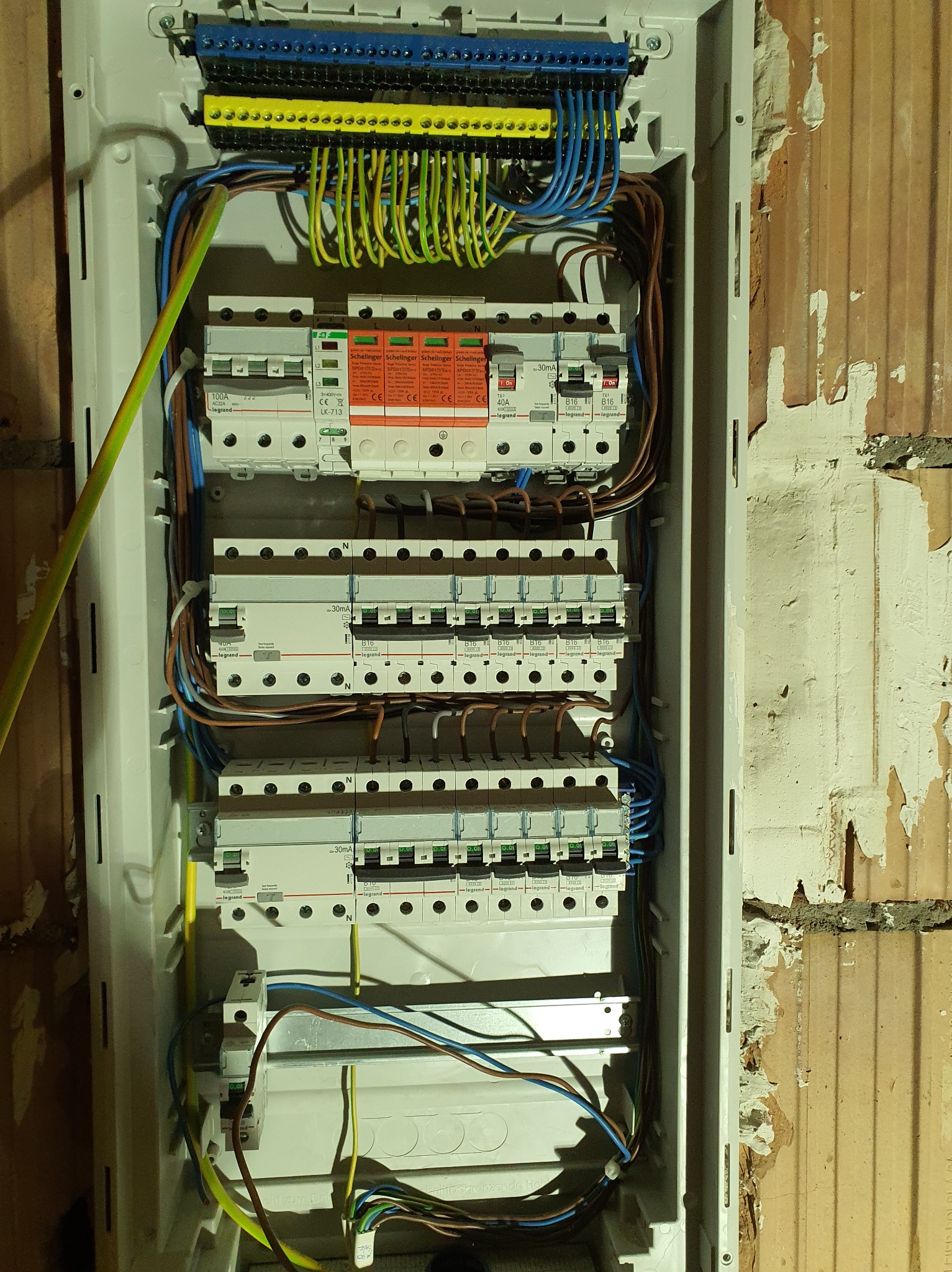 Rozdzielnica elektryczna Legrand z zabezpieczeniami przepięciowymi Schellinger, zamontowana na ścianie z cegły. Widoczne kable i bezpieczniki.