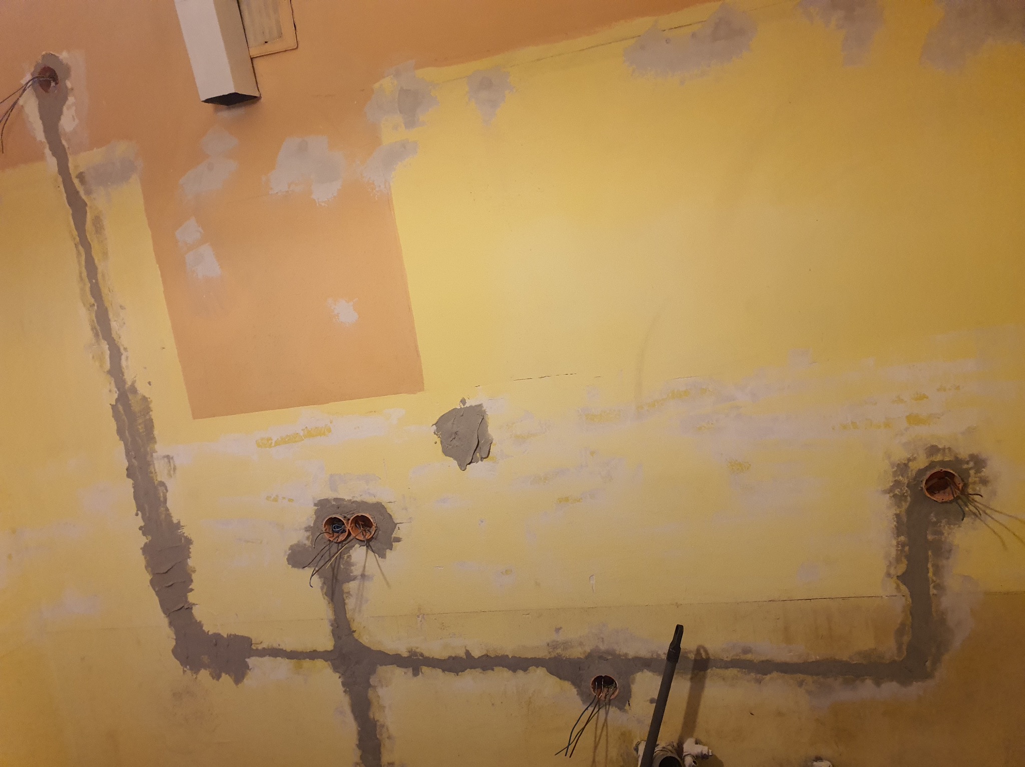 Żółta ściana w trakcie remontu z widocznymi puszkami instalacyjnymi i bruzdami wypełnionymi gipsem, przygotowana pod dalsze prace wykończeniowe.