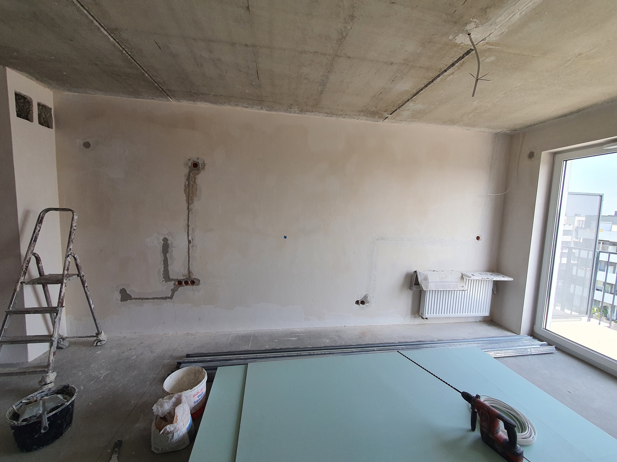 Surowy stan wnętrza z widocznymi przewodami, puszkami elektrycznymi w ścianie, narzędziami i materiałami budowlanymi. W tle okno z widokiem na budynek.