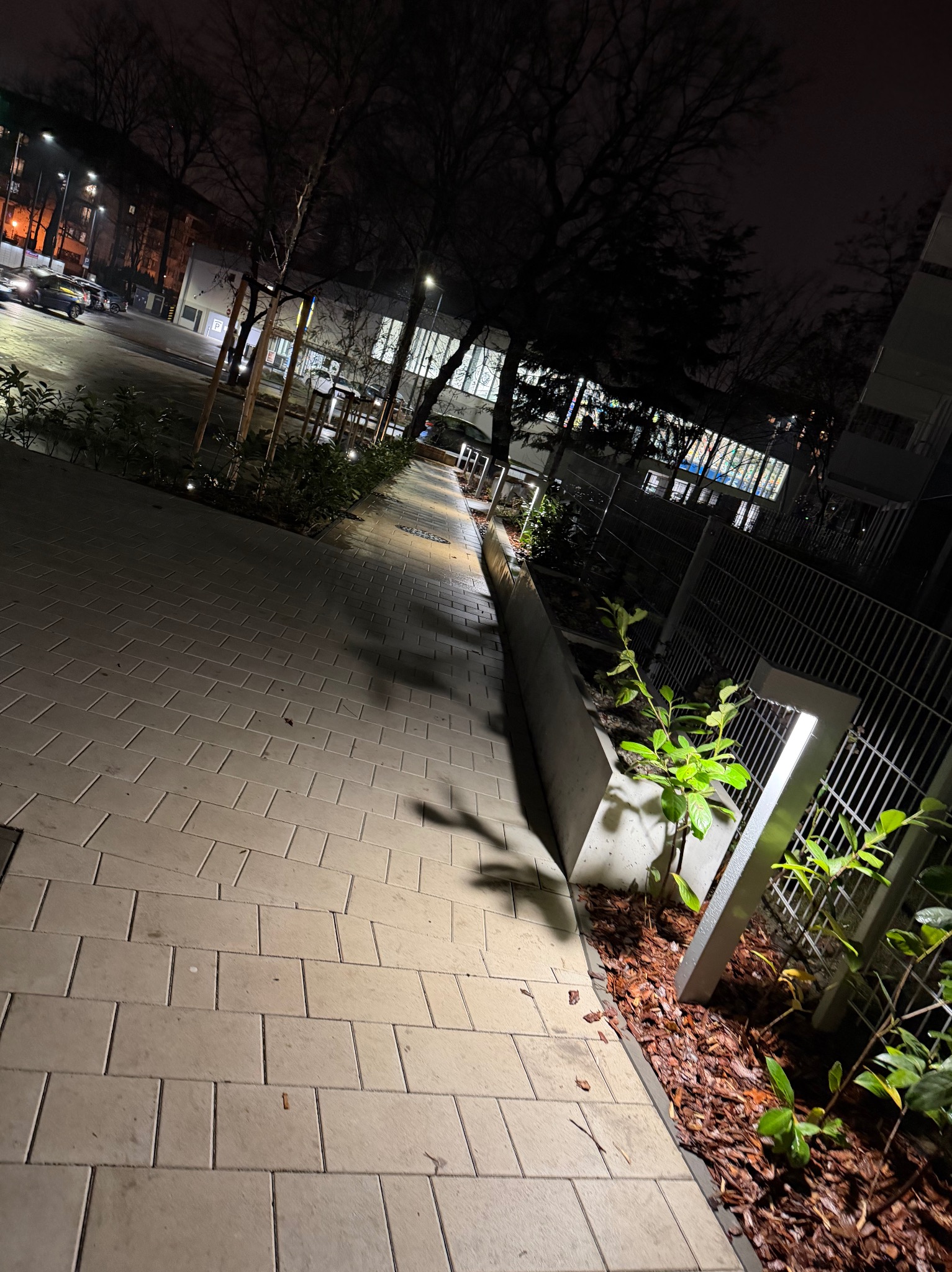 Nocne oświetlenie ścieżki z kostki brukowej, z lampami wzdłuż ogrodzenia i roślinności. Widok na budynek w tle, oświetlony ulicznymi latarniami.