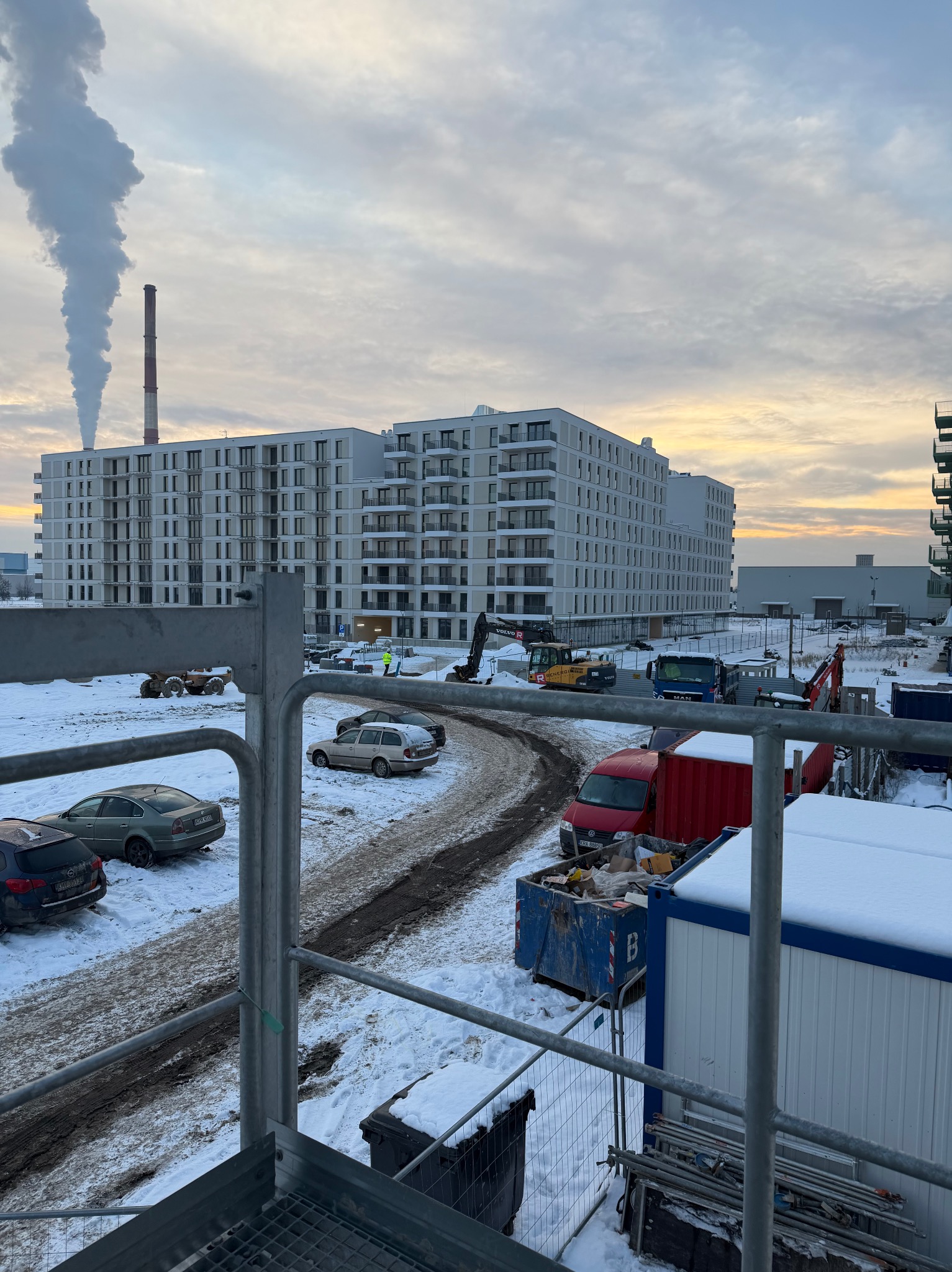 Zimowy krajobraz budowy z blokami mieszkalnymi w tle, widok z góry przez metalową konstrukcję. Na pierwszym planie kontenery i samochody, w oddali komin z dymem.