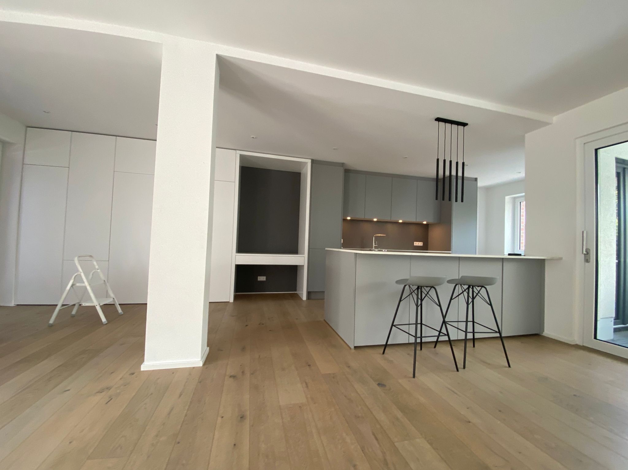 Jasne, przestronne wnętrze po remoncie: drewniana podłoga, szara kuchnia z wyspą i stołkami, białe ściany i szafy. Drabina malarska w rogu sugeruje świeże malowanie.