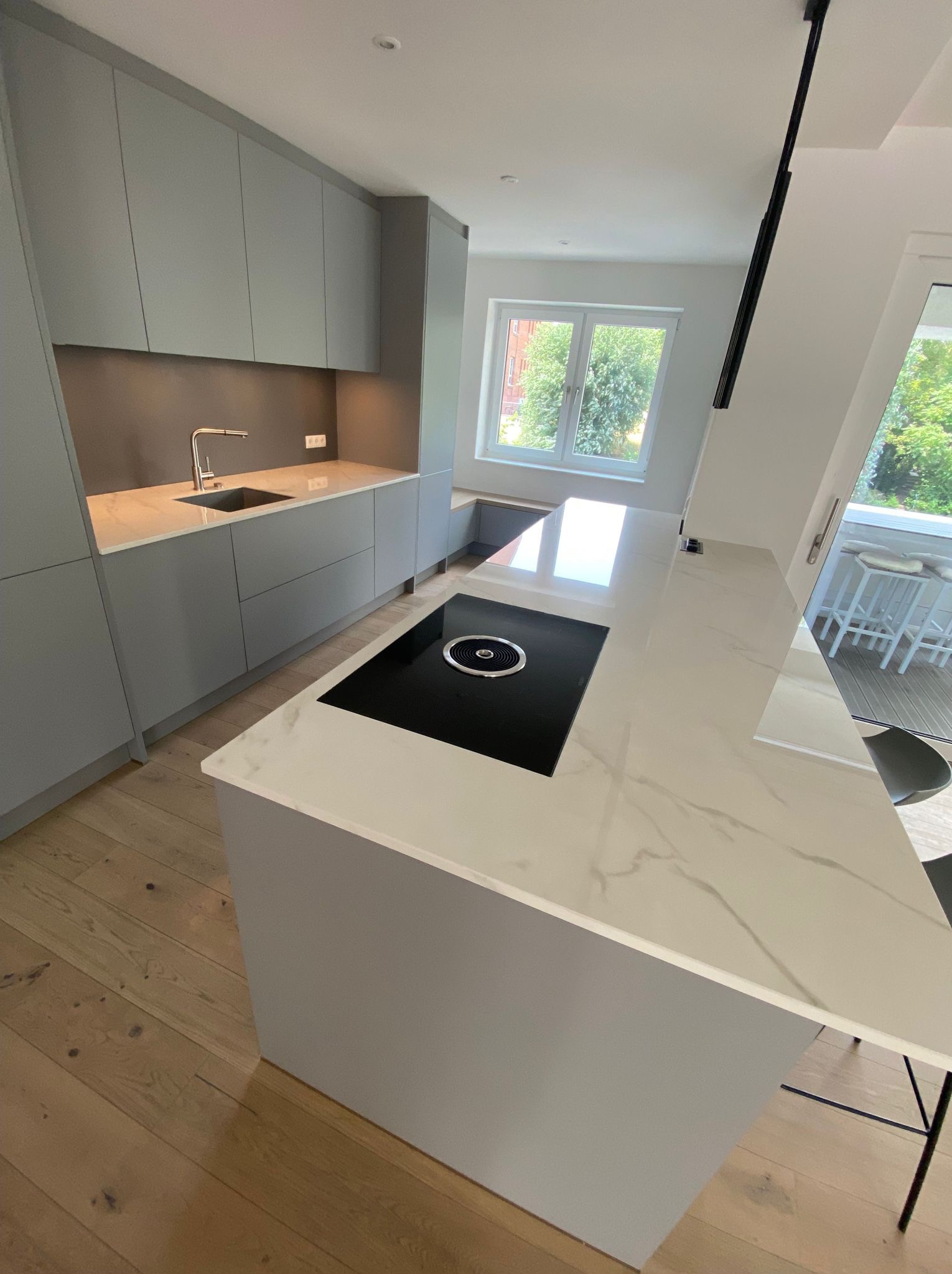 Nowoczesna, szara kuchnia z marmurowym blatem i płytą indukcyjną. Widok na okno i balkon. Jasne drewniane podłogi. Minimalistyczny design.