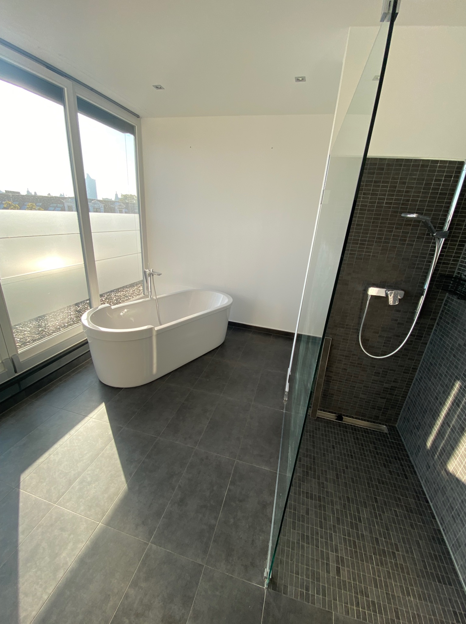 Nowoczesna łazienka z wolnostojącą wanną przy oknie i kabiną prysznicową z ciemną mozaiką. Minimalistyczny design, szare płytki podłogowe, jasne światło słoneczne.
