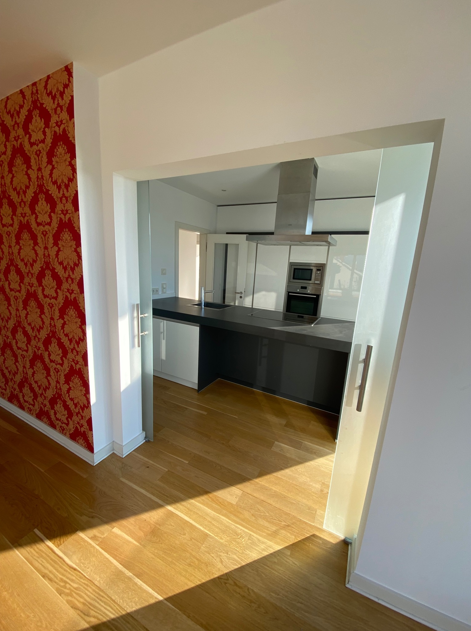 Nowoczesna kuchnia z białymi szafkami i ciemnym blatem, widziana z perspektywy pokoju z wzorzystą, czerwoną tapetą. Parkiet w ciepłym odcieniu drewna.