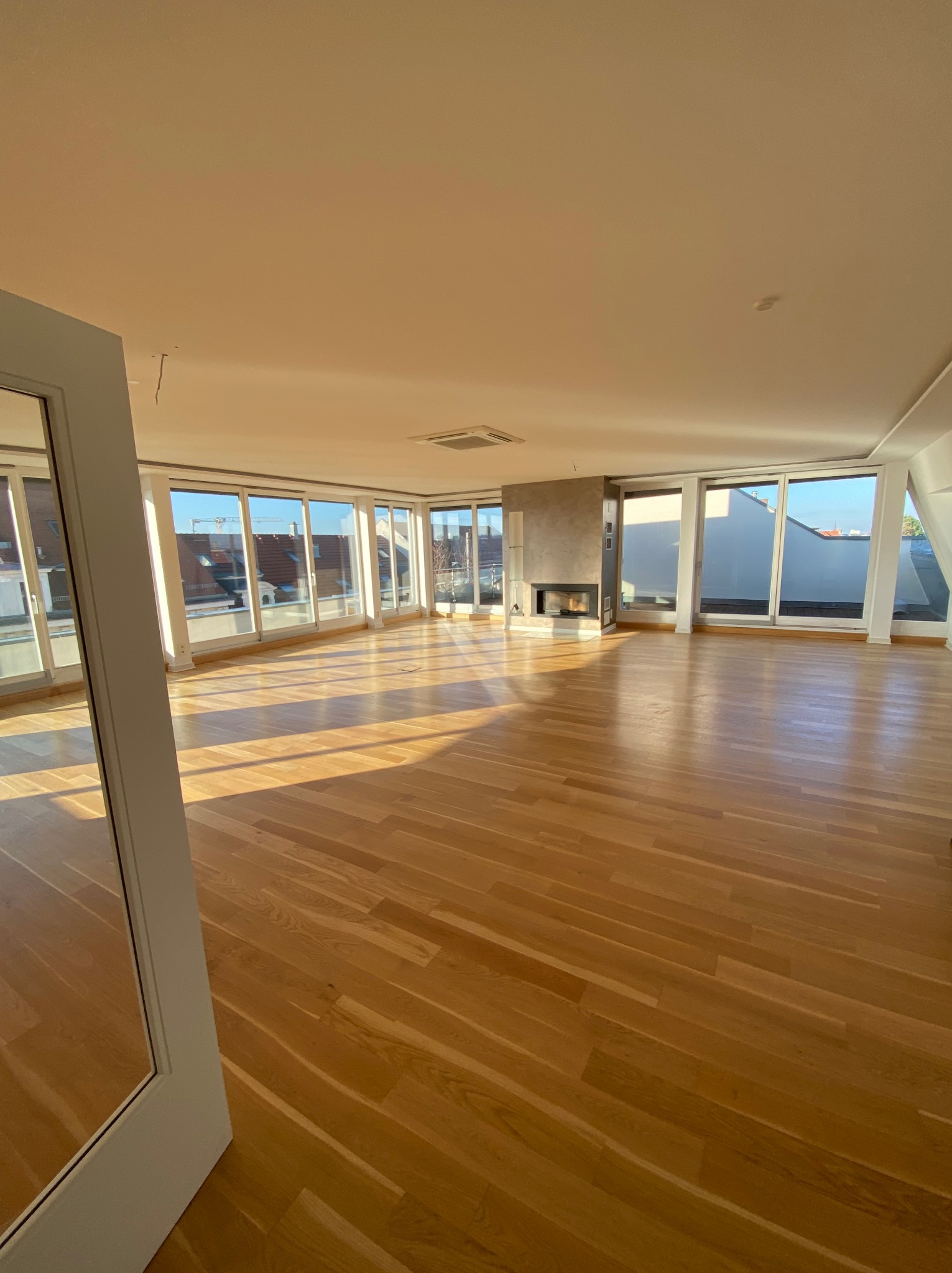 Jasne, przestronne wnętrze apartamentu z parkietem, kominkiem i panoramicznymi oknami. Widok na miasto zza okien. Częściowo widoczne drzwi z lustrem po lewej.