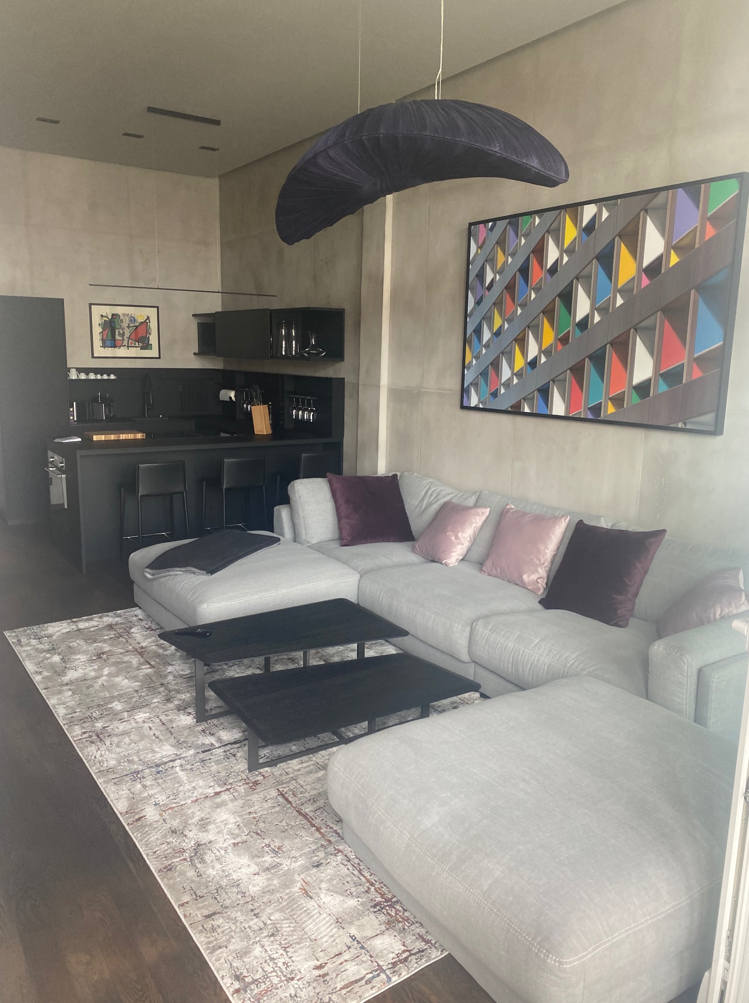 Szary narożnik z poduszkami w salonie z aneksem kuchennym, nad nim designerska lampa i obraz z kolorowymi trójkątami. Nowoczesne wnętrze po sprzątaniu.