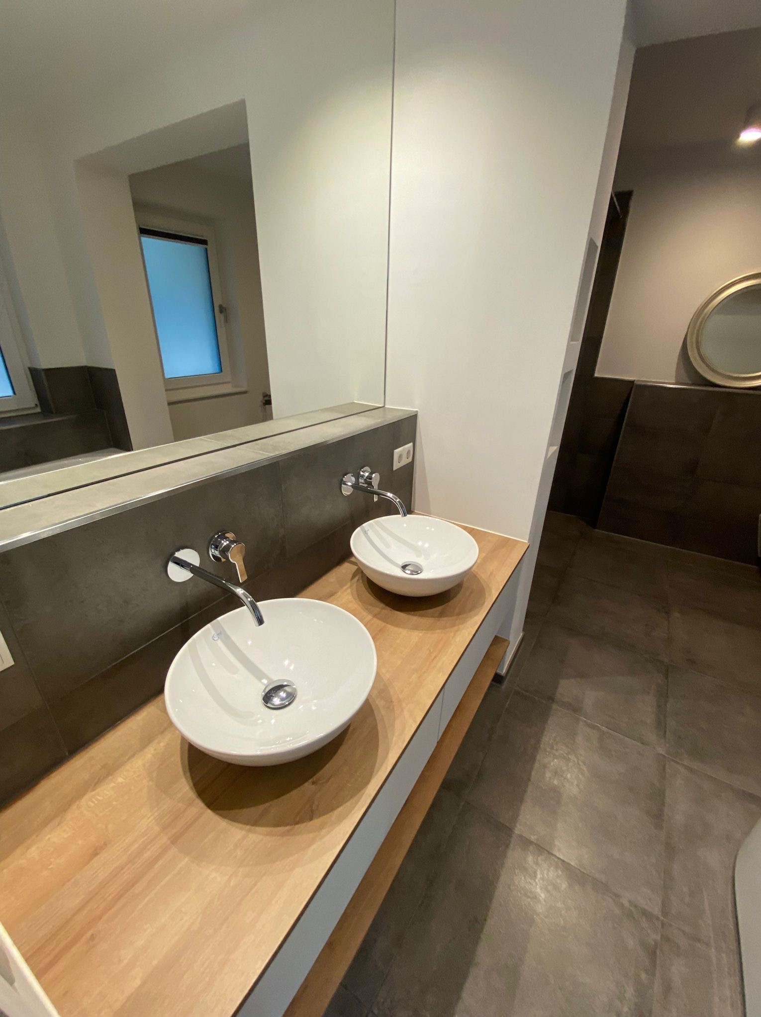 Nowoczesna łazienka z dwiema okrągłymi umywalkami nablatowymi na drewnianym blacie, szara glazura i duże lustro. Widok na fragment podłogi i lustro w tle.