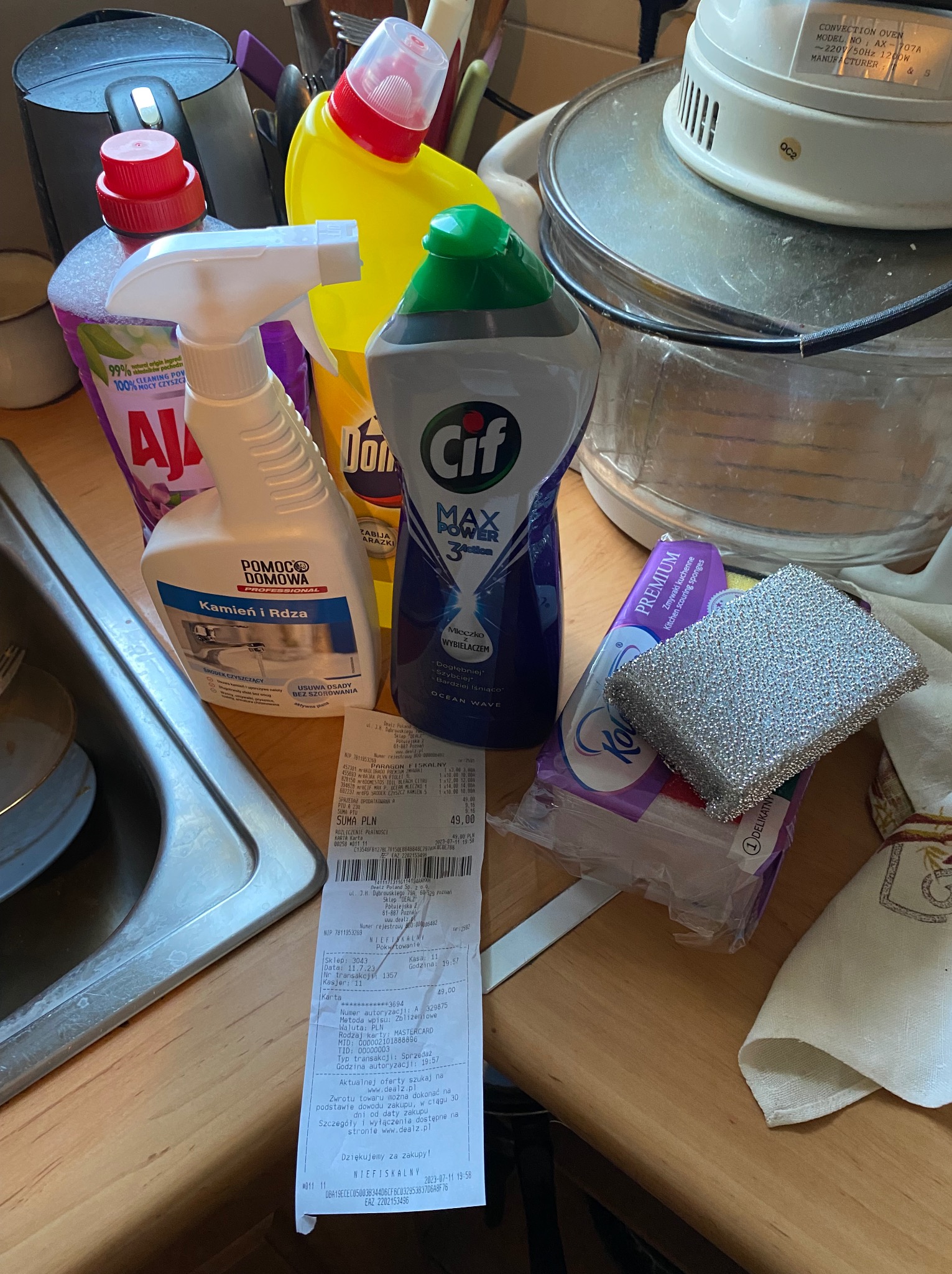 Środki czystości: Cif, Domestos, Ajax, Pomoc Domowa, zmywak, paragon na blacie kuchennym obok zlewu i piekarnika konwekcyjnego. Artykuły do sprzątania.