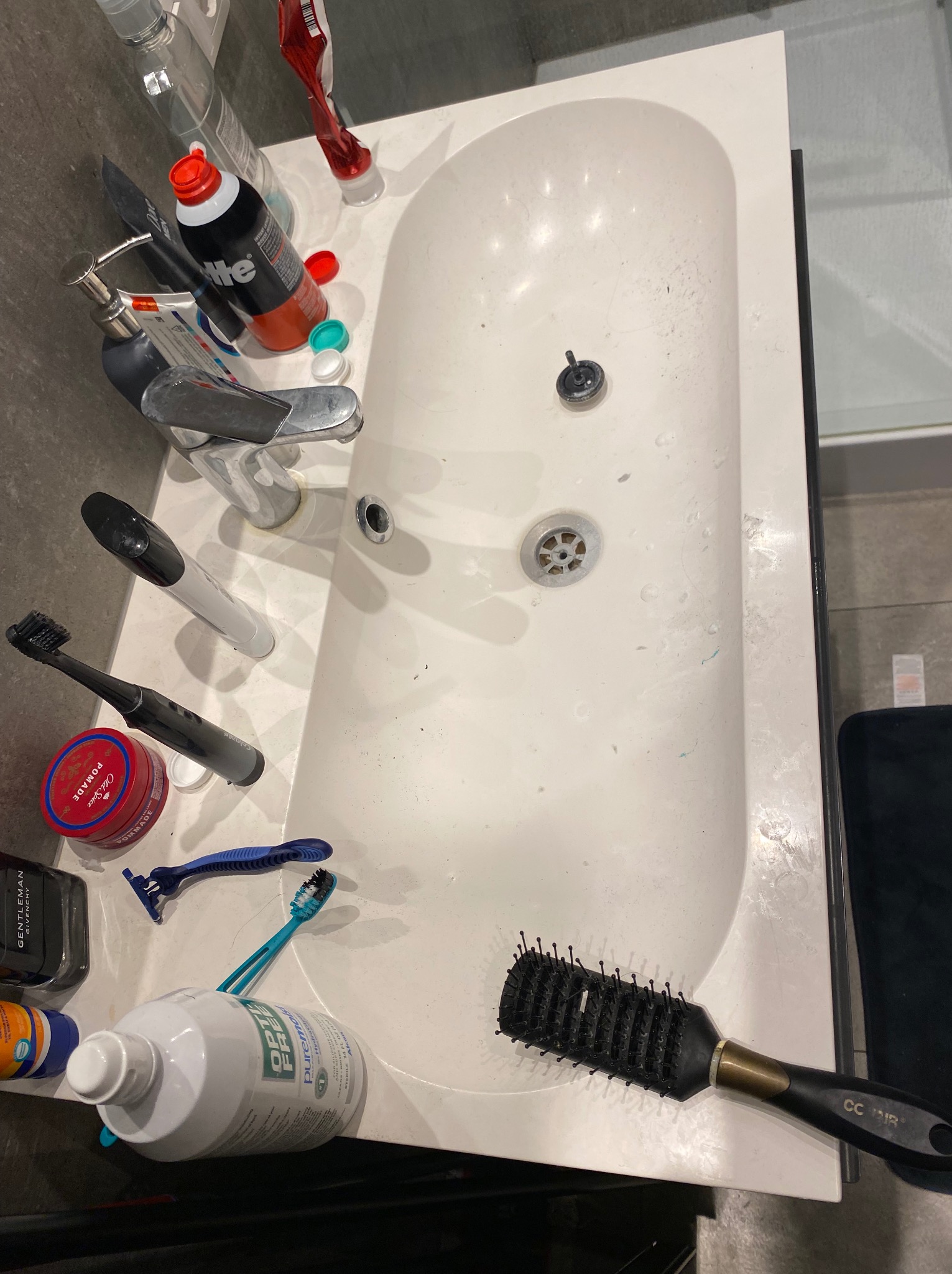 Zabrudzona umywalka z kosmetykami, szczoteczkami i przyborami toaletowymi. Widoczne ślady użytkowania i drobne zabrudzenia na powierzchni umywalki.