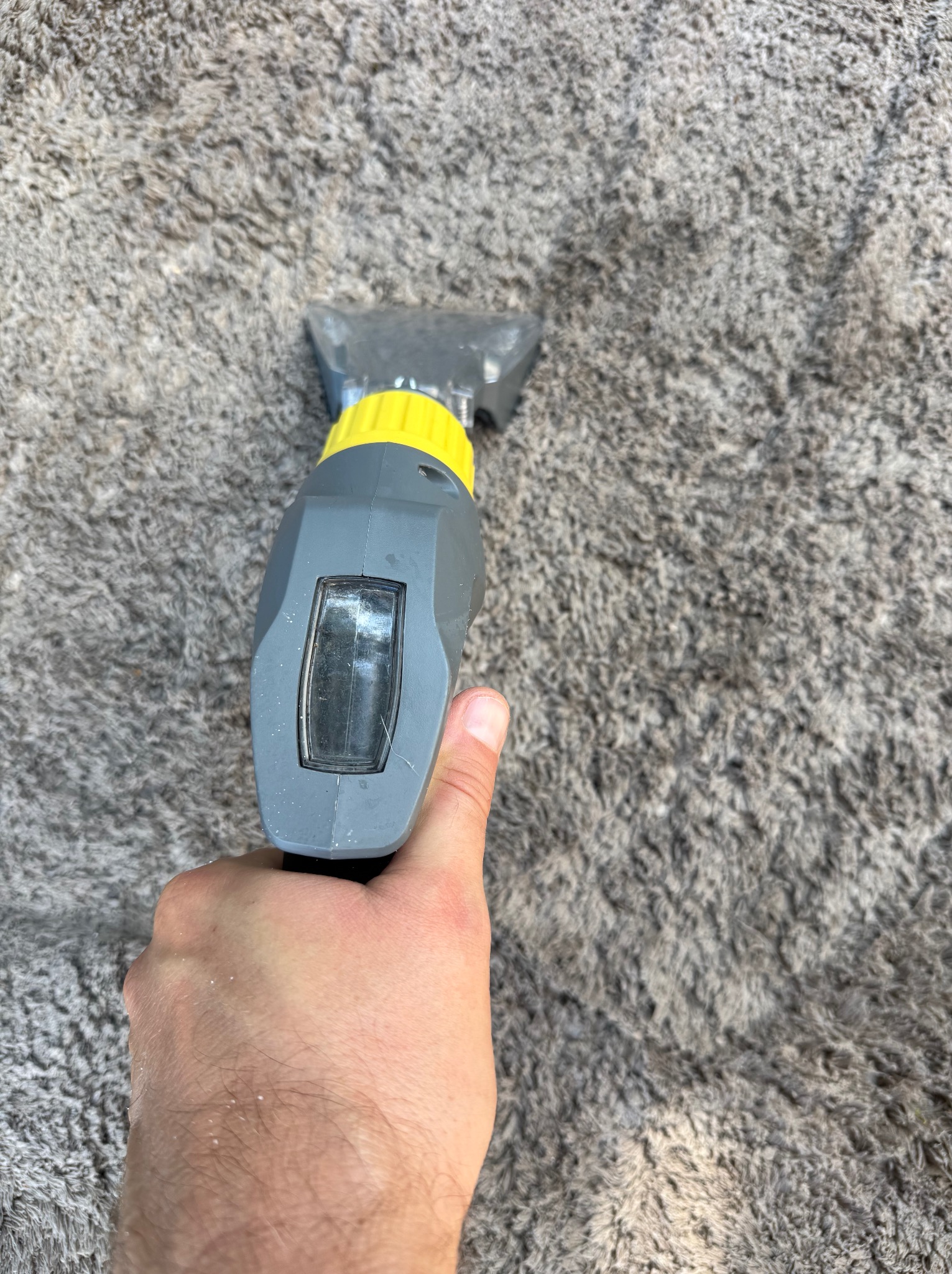 Czyszczenie dywanu szarym odkurzaczem piorącym trzymanym w dłoni, widok z góry na fragment dywanu z długim włosiem podczas czyszczenia.