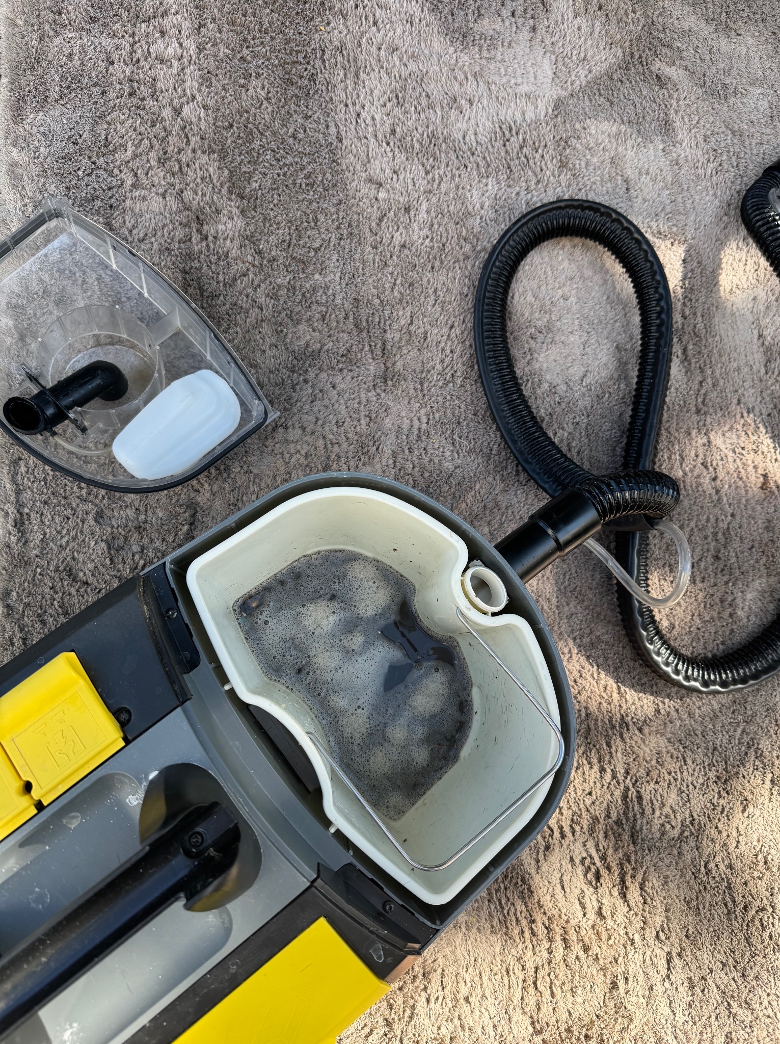 Zbliżenie na zbiornik z brudną wodą i pianą z odkurzacza piorącego, leżącego na dywanie. Widoczny wąż i pojemnik na czystą wodę. Sprzęt do prania dywanów.