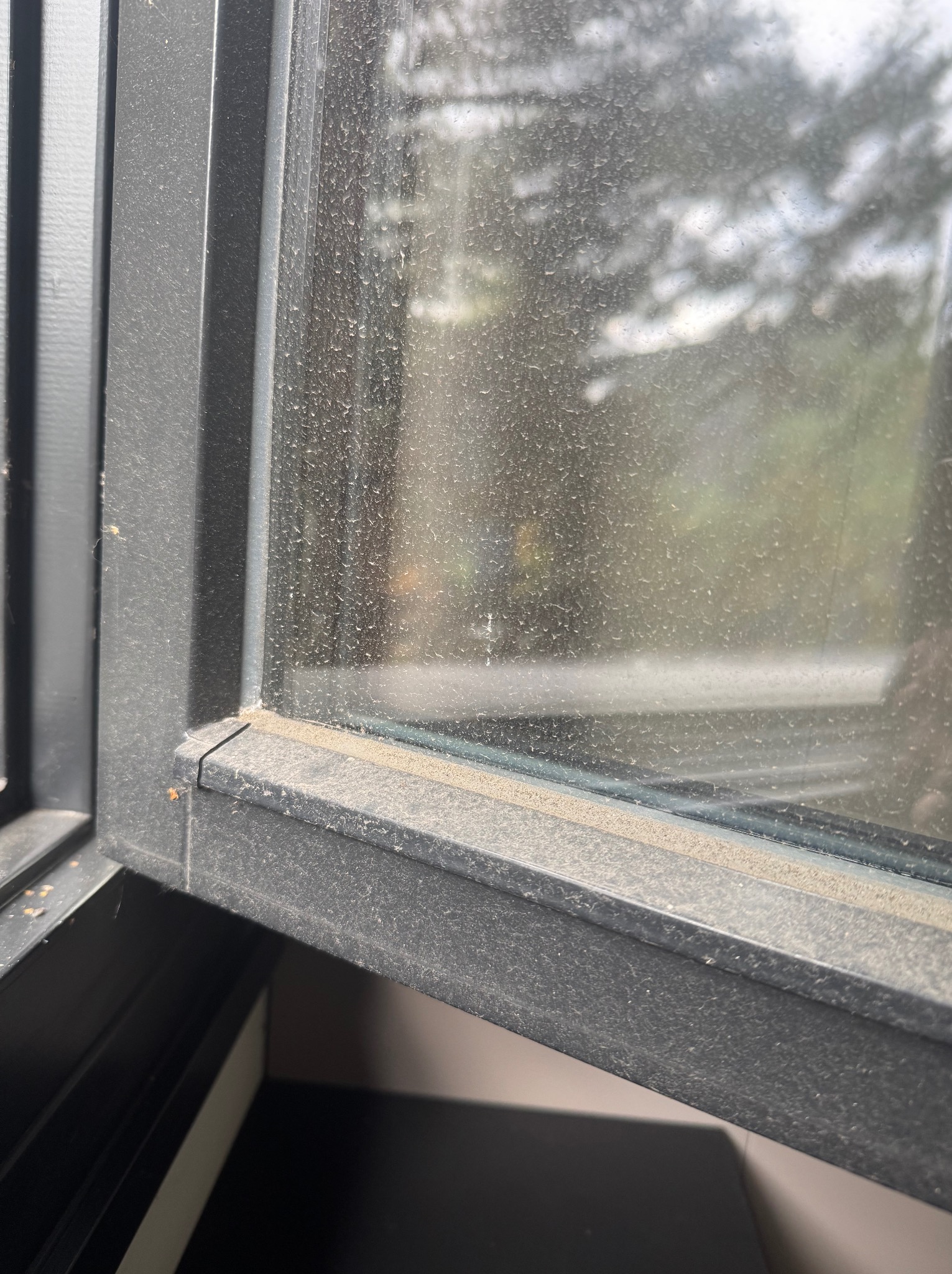 Bardzo brudne okno z szarą ramą, pokryte grubą warstwą kurzu i zabrudzeń, z widocznymi smugami i zaciekami. Widok na zewnątrz jest mocno zamglony.