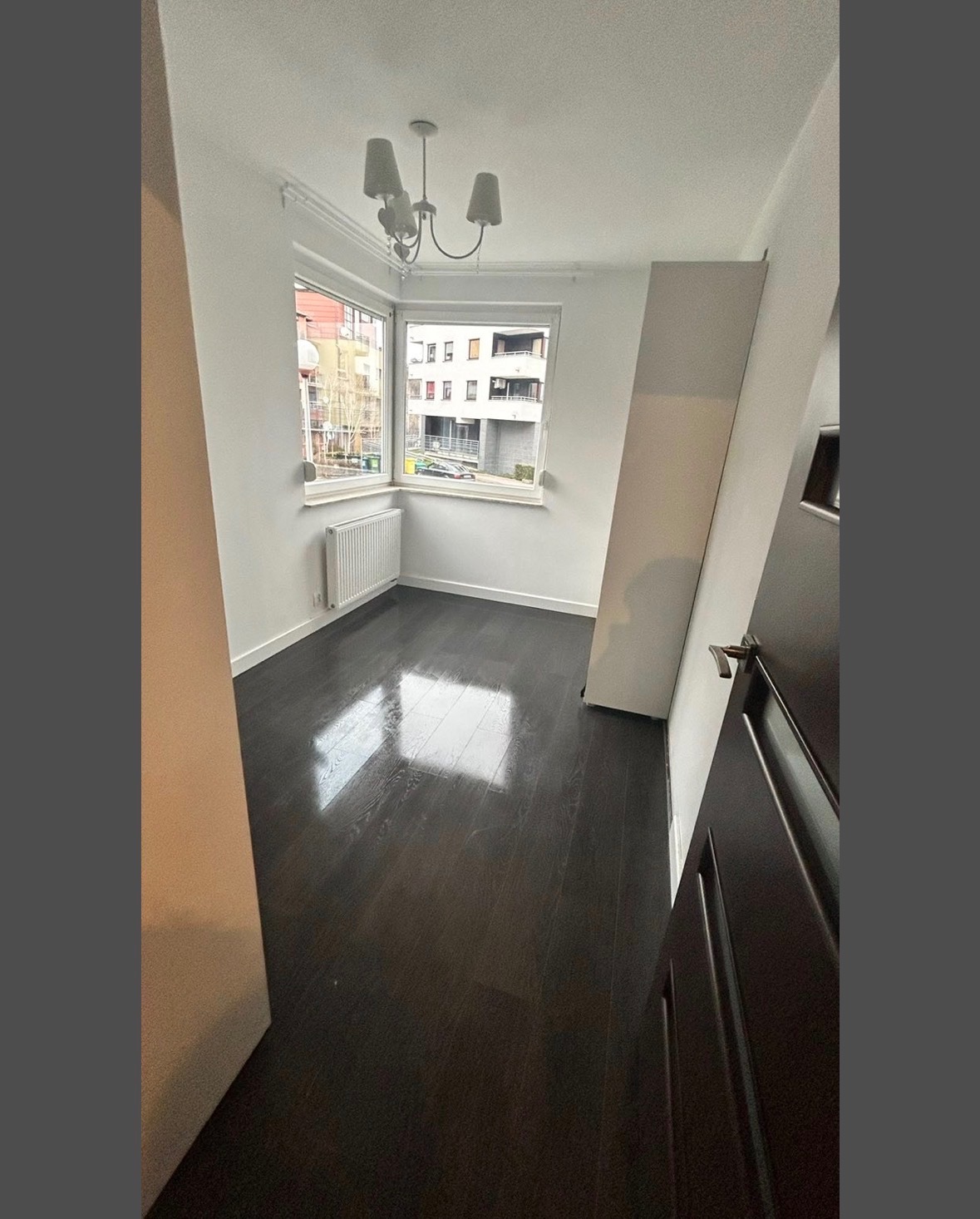 Pusty pokój po sprzątaniu: lśniąca, ciemna podłoga odbija światło z okna. Białe ściany, grzejnik i widok na budynek zza okna. Czystość i porządek.