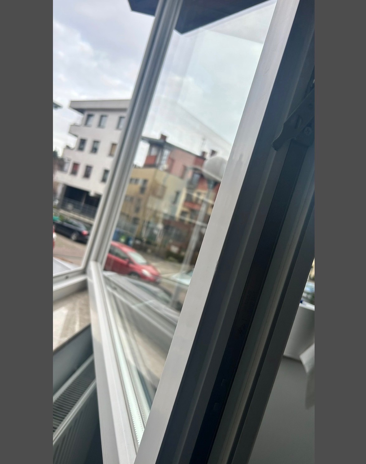 Białe, otwarte okno z widokiem na budynki mieszkalne i samochody w pochmurny dzień. Ujęcie z bliska, z ostrym kątem, ukazujące detale ramy okiennej.
