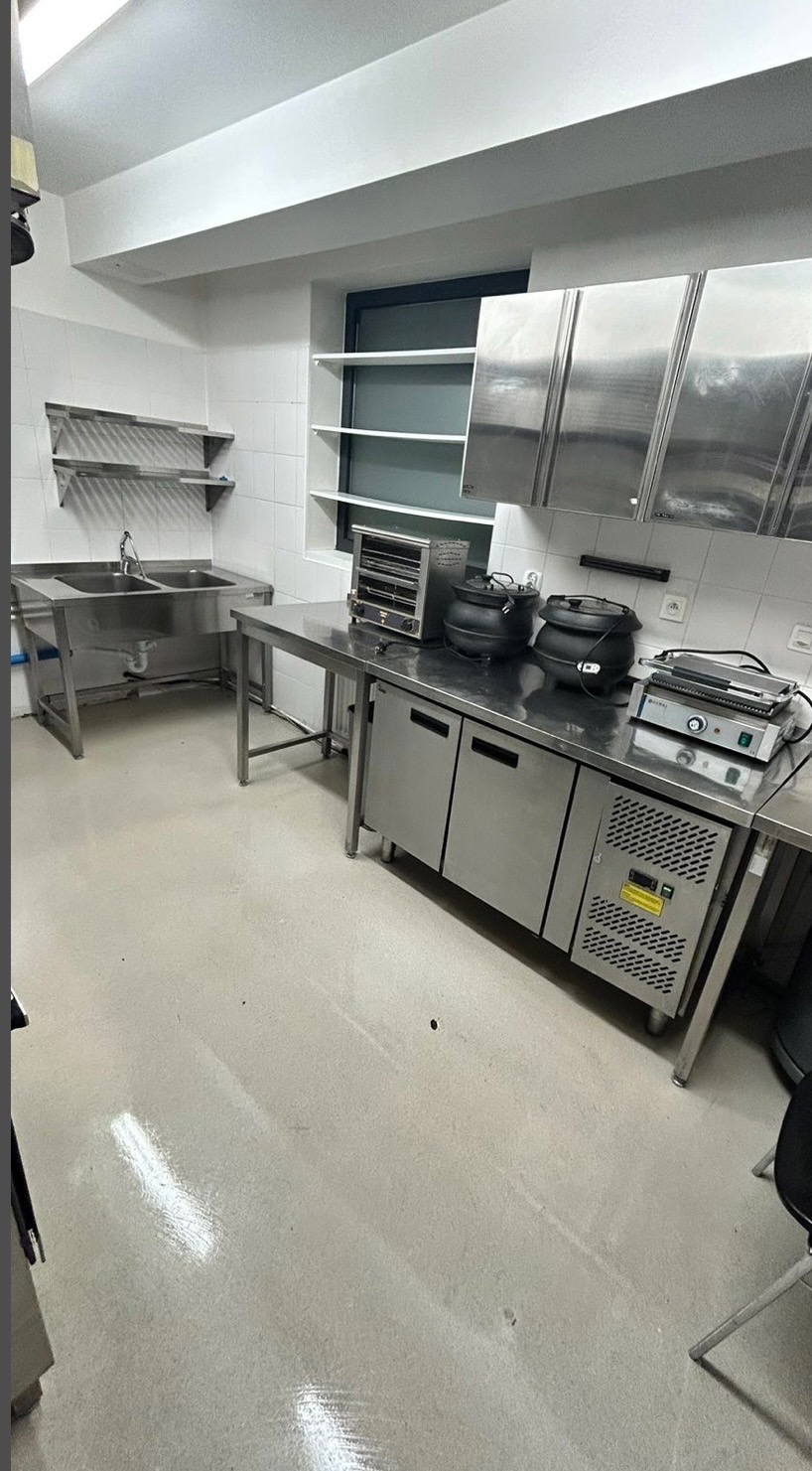 Wnętrze kuchni gastronomicznej ze stali nierdzewnej: zlewozmywak, szafki chłodnicze, półki i sprzęt kuchenny. Białe płytki na ścianach, jasna podłoga.