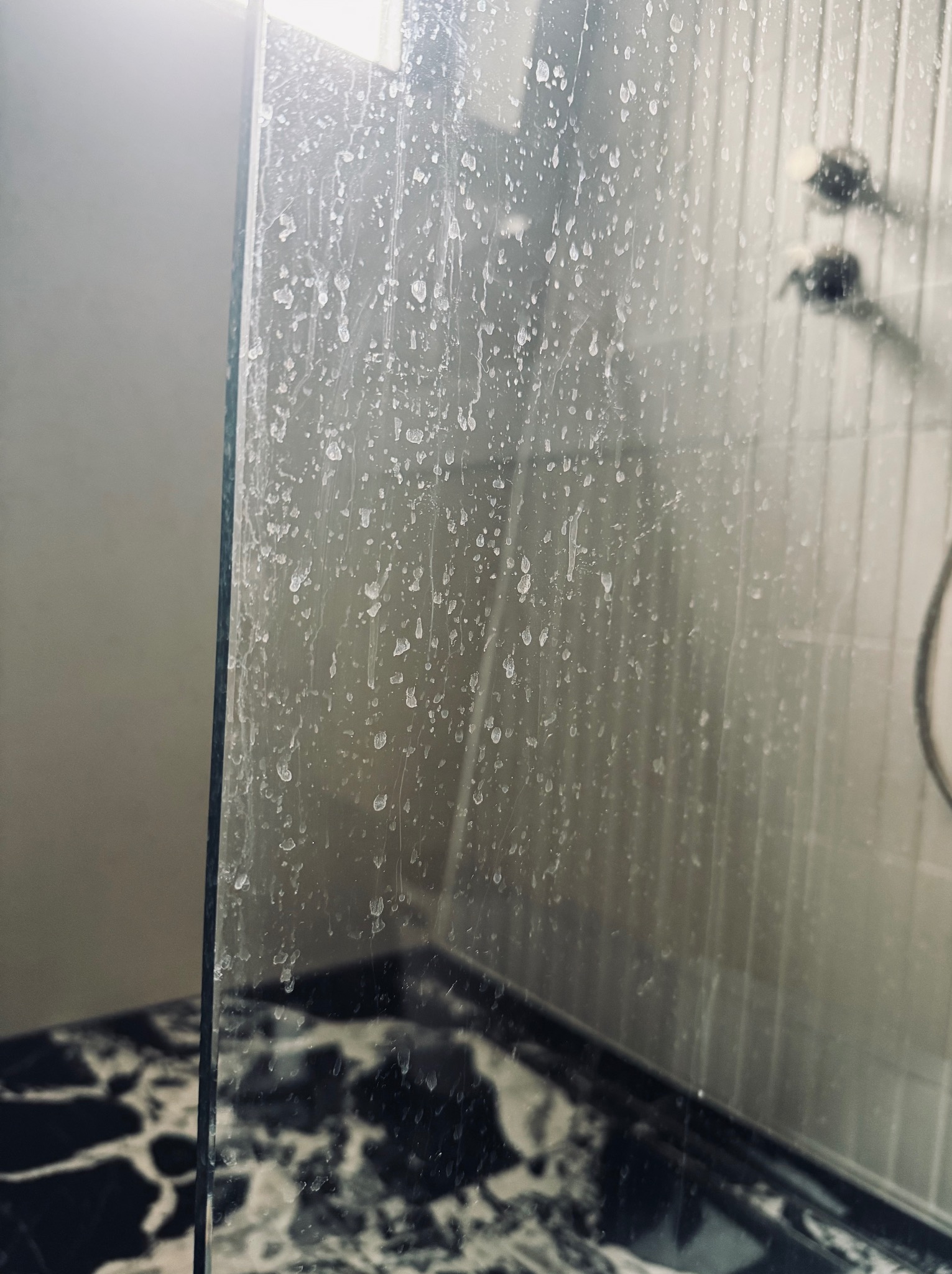 Z bliska: Zakamieniona szyba kabiny prysznicowej z widocznymi zaciekami i osadami, na tle płytek i armatury. Potrzeba gruntownego czyszczenia.