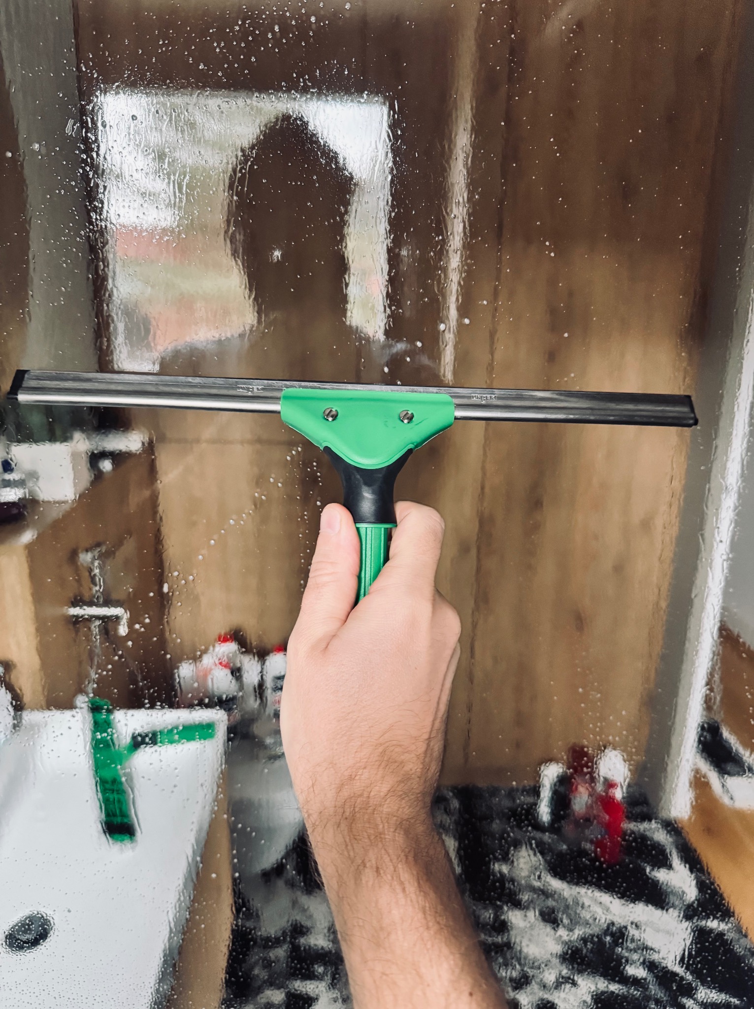Czyszczenie szyby prysznicowej z zaciekami za pomocą ściągaczki z zielonym uchwytem. Widoczna umywalka i fragment łazienki w tle.
