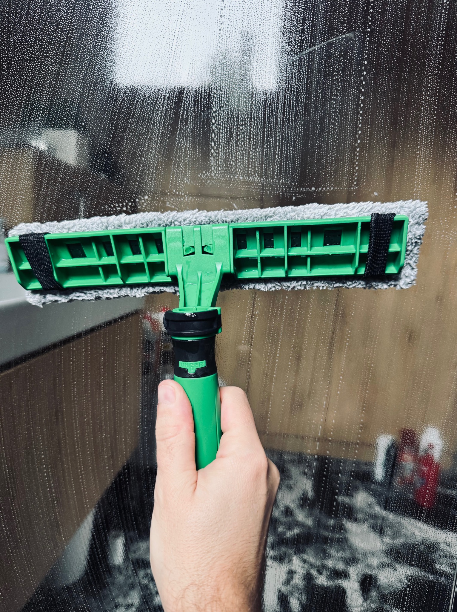 Mycie okna z użyciem zielonej myjki z szarą nakładką, widoczne smugi piany na szybie i odbicie wnętrza pomieszczenia. Dłoń trzyma narzędzie.