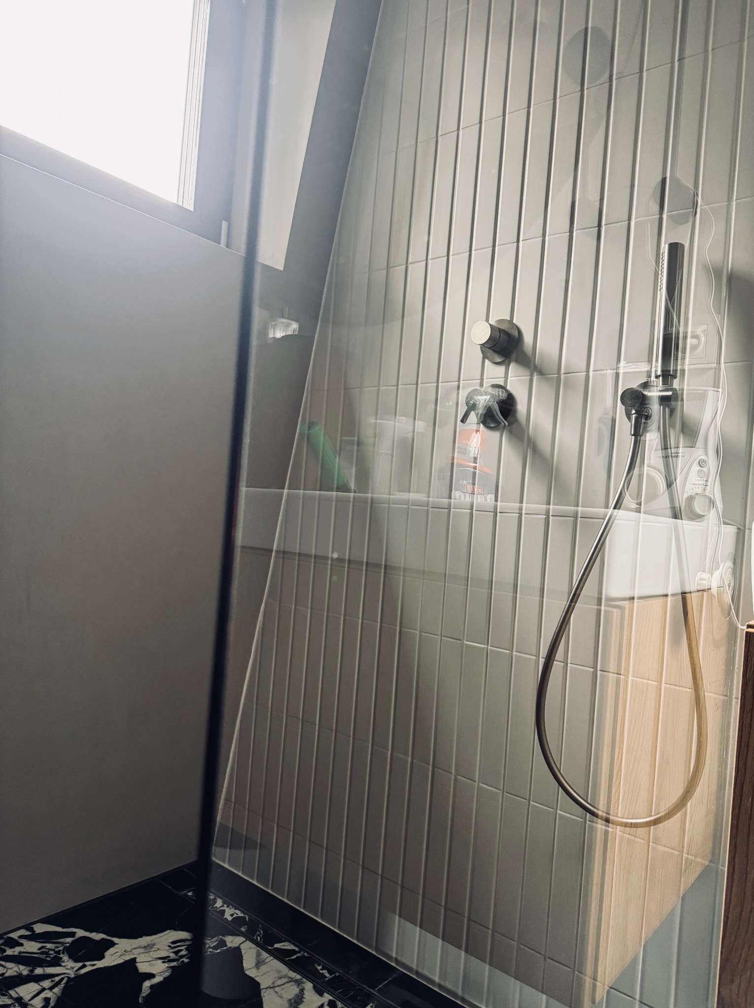 Nowoczesna kabina prysznicowa z szarymi, pionowymi płytkami, czarną marmurową podłogą i szklaną ścianką. Widoczne elementy armatury i akcesoria łazienkowe.