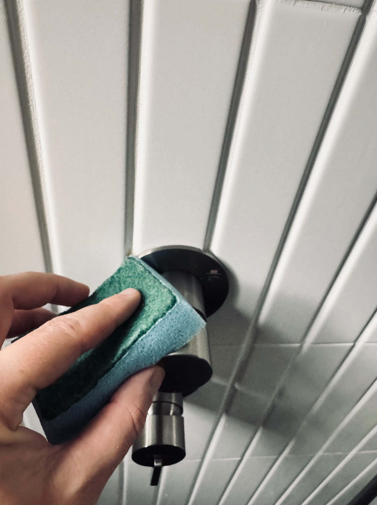 Czyszczenie chromowanej armatury łazienkowej gąbką na tle białych, wypukłych płytek ściennych. Ujęcie z dołu, skupione na detalu.