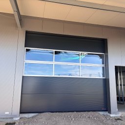 Dobre bramy - Ciemnoszara brama segmentowa z oknami w hali przemysłowej, widoczny fragment konstrukcji budynku i surowy teren przed budynkiem. Brama marki Hormann.