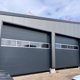 Dobre bramy - Dwie szare bramy garażowe segmentowe z oknami, zamontowane w szarym budynku przemysłowym. Widoczna konstrukcja budynku i fragmenty pojazdów.