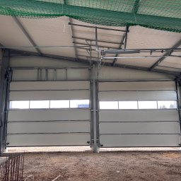 Dobre bramy - Dwie segmentowe bramy przemysłowe z oknami, częściowo otwarte, w surowym wnętrzu hali z widoczną konstrukcją dachu i elementami instalacji. Widoczny piasek na podłodze.