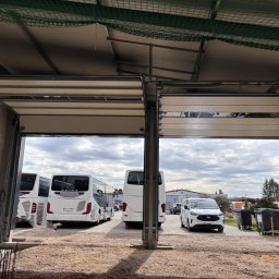 Dobre bramy - Otwarta brama przemysłowa z widokiem na autobusy i samochód osobowy na zewnątrz. Widoczna konstrukcja bramy i fragment dachu z siatką zabezpieczającą.