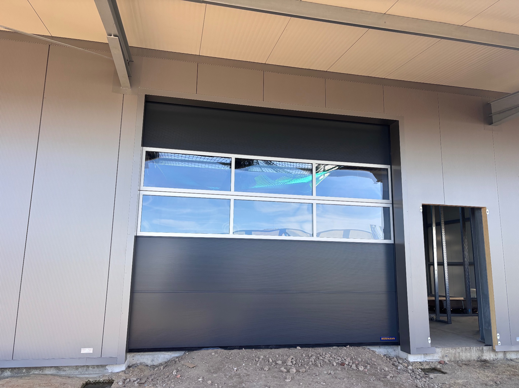 Ciemnoszara brama segmentowa z oknami w hali przemysłowej, widoczny fragment konstrukcji budynku i surowy teren przed budynkiem. Brama marki Hormann.