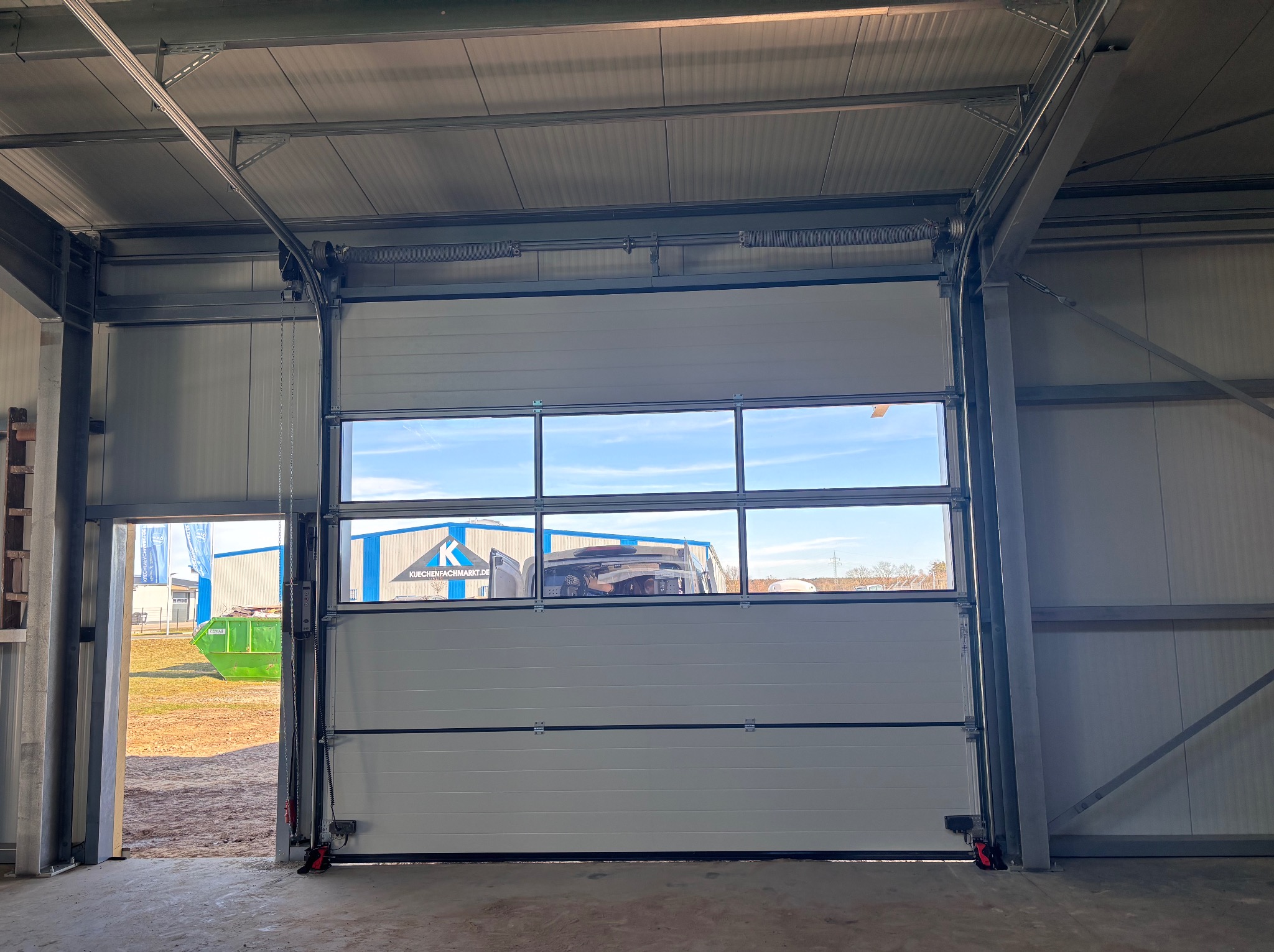 Otwarta segmentowa brama garażowa z oknami, widok z wnętrza hali. Na zewnątrz kontener i budynek z logo firmy. Widoczne elementy konstrukcyjne bramy.