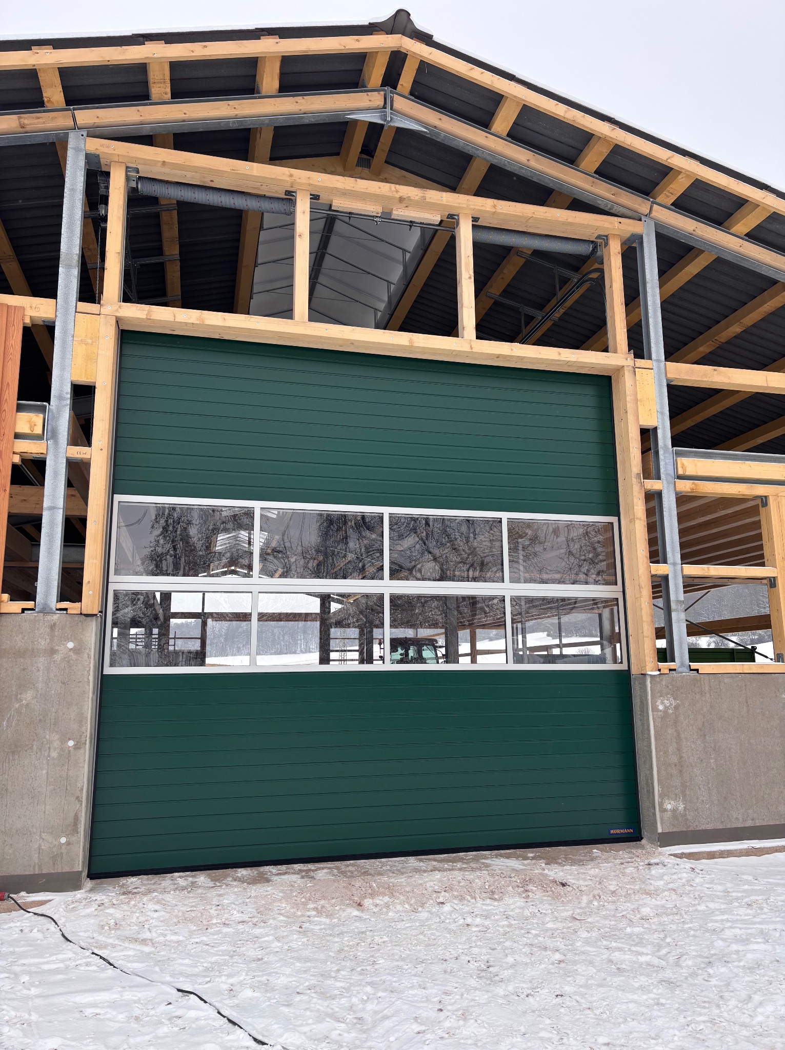 Zielona brama segmentowa z oknami w surowej konstrukcji budynku gospodarczego, widok z zewnątrz na tle zimowego krajobrazu. Widoczne elementy konstrukcyjne.