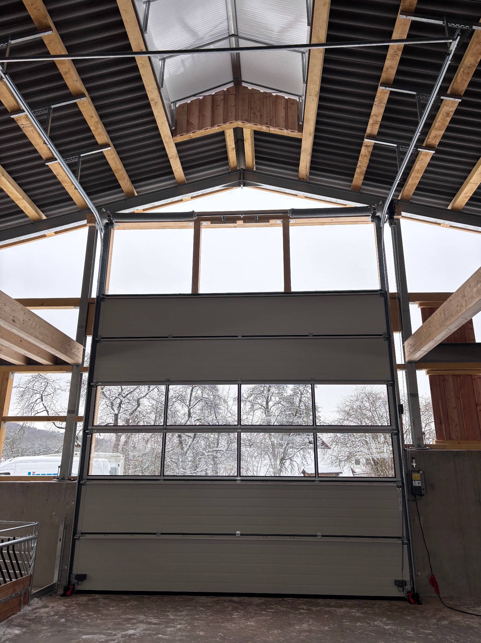 Segmentowa brama garażowa z oknami, widok od wewnątrz, konstrukcja dachu z drewnianymi belkami, zimowy krajobraz za oknami bramy, częściowo otwarta.