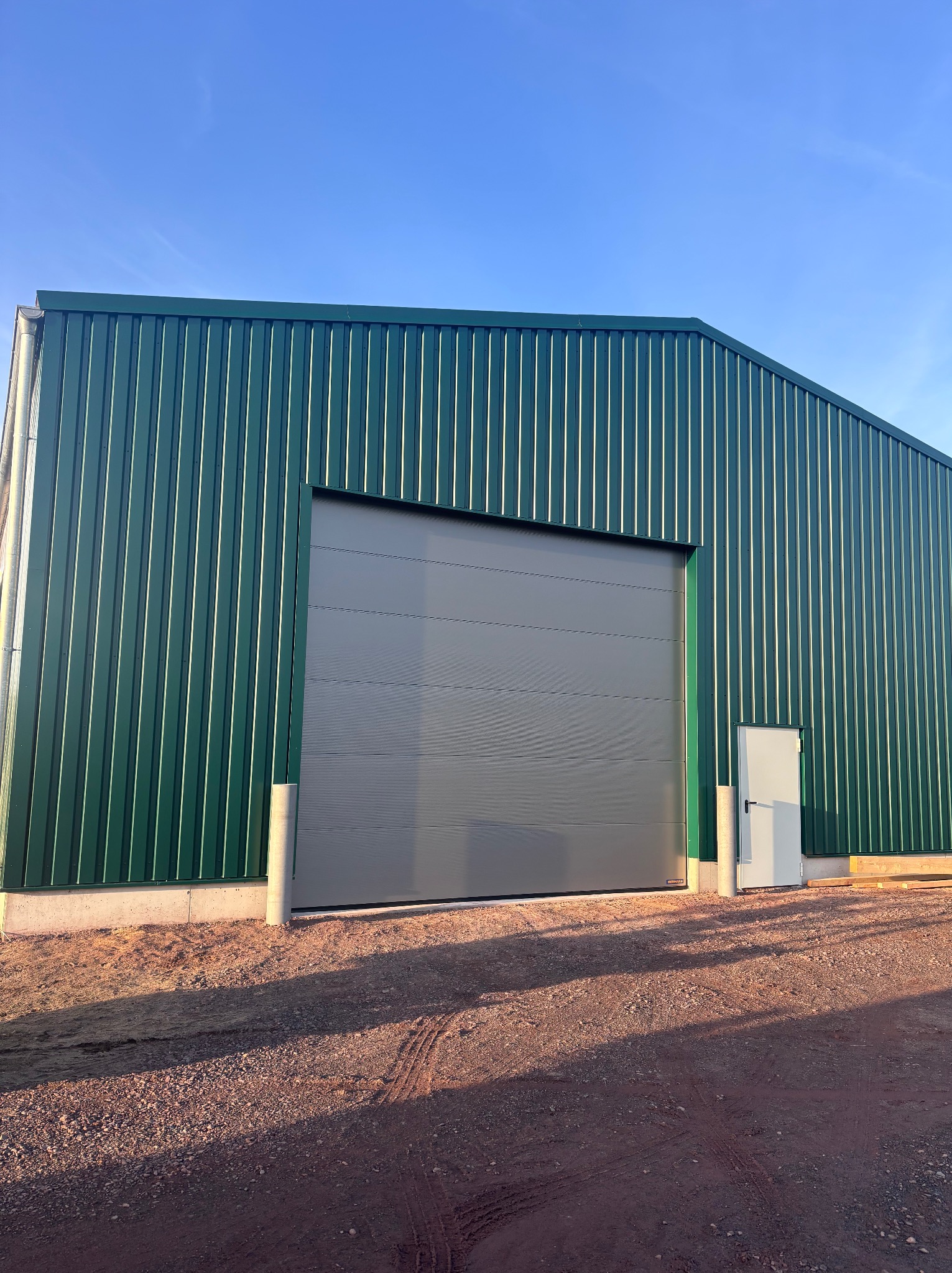 Zielony budynek przemysłowy z szarą bramą segmentową i białymi drzwiami wejściowymi. Widok z zewnątrz, w słoneczny dzień.